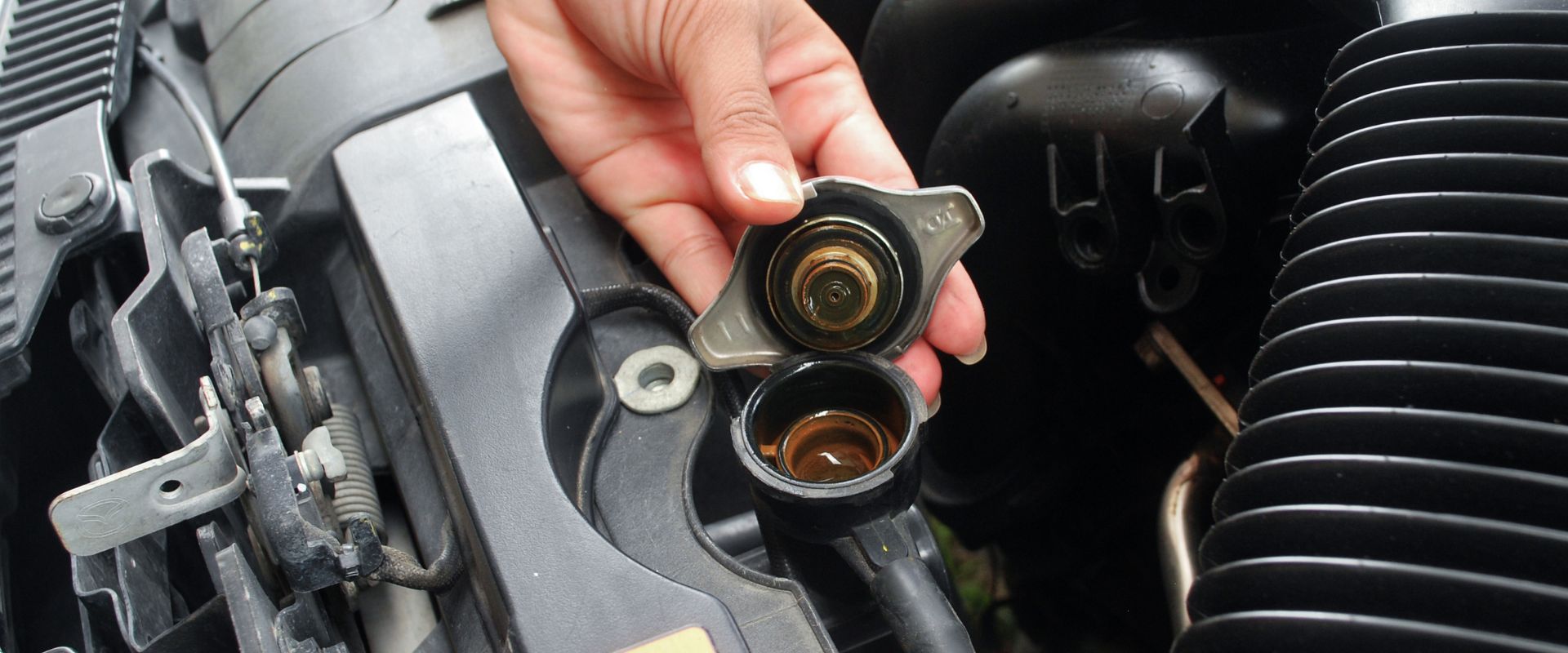 Hand holding a car radiator cap above the engine, illustrating vehicle maintenance and cooling system inspection for auto care services in Dallas, TX.